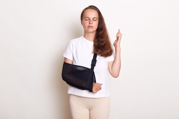 Unhealthy arm broken Caucasian woman with arm sling supported on her hand being injured keeps eyes closed and crossed fingers having hope for fast recovery posing isolated on white background