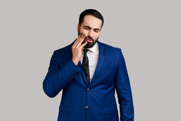 Unhealthy bearded man wincing in pain and touching sore cheek, suffering unbearable toothache, gum disease, wearing official style suit. Indoor studio shot isolated on gray background.