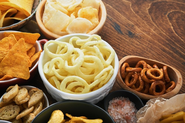 Unhealthy food or snacks All classic potato snacks with peanuts popcorn and onion rings and salted pretzels in bowl plates on old wooden background Unhealthy food for figure heart skin teeth