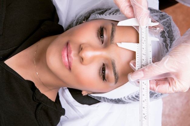 Unidentified beautician in gloves gown measures the proportions of the eyebrows