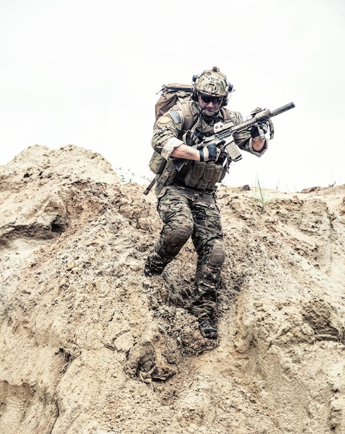 United States army commando, special forces infantry armed with assault rifle in combat uniform and load carrier, descending from steep sand dune. Armed conflict in desert, soldier rush across dunes