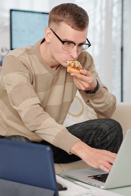 University student in glasses eating slice of pizza when working on laptop at home