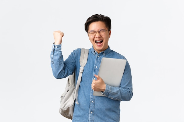 University, study abroad and lifestyle concept. Happy rejoicing asian male student with braces triumphing, pass exams, finish final semester, fist pump and shouting yes with satisfaction, hold laptop