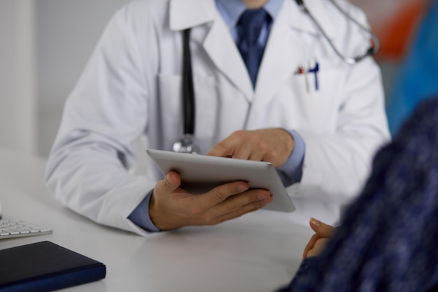 Unknown bearded doctor and patient woman discussing current health examination while sitting and using tablet computer, close-up. Medicine concept.