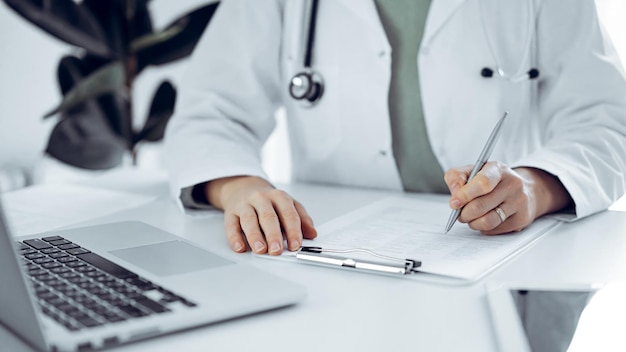 Unknown doctor woman sitting and writing notes at the desk in clinic or hospital office, close up. Medicine concept.