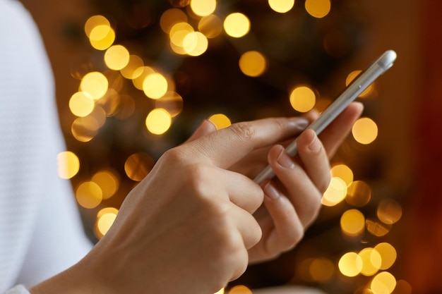 Unknown female hand holding mobile phone and typing texting with friends browsing Internet anonymous woman posing with smart phone with blurred Christmas tree on background