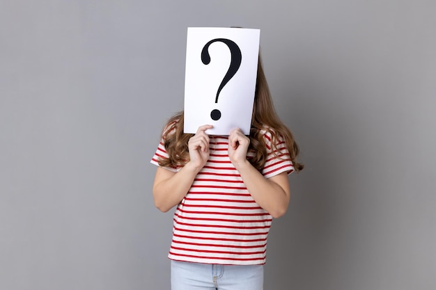 Unknown little girl wearing striped Tshirt standing hiding face behind paper with question mark