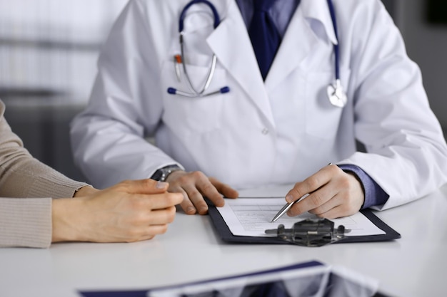 Unknown male doctor and patient woman discussing something while sitting in clinic and using clipboard. Best medical service in hospital, medicine, pandemic stop.