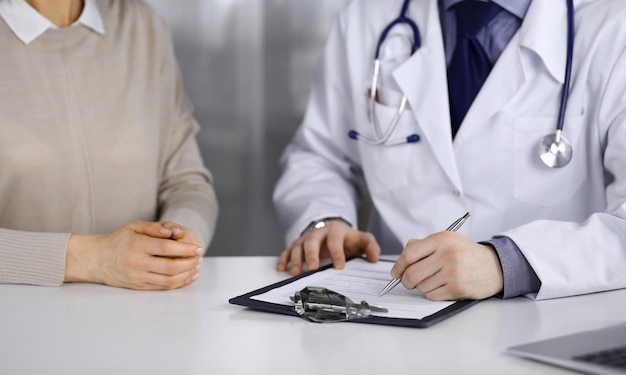 Unknown male doctor and patient woman discussing something while sitting in clinic and using clipboard. Best medical service in hospital, medicine, pandemic stop.