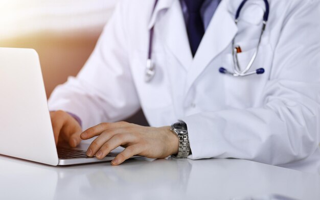 Unknown male doctor sitting and working with laptop in a darkened clinic, glare of light on the background, close-upof hands.