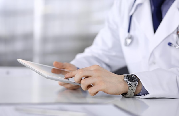 Unknown male doctor sitting and working with tablet computer in clinic at his working place, close-up. Young physician at work. Perfect medical service, medicine concept.