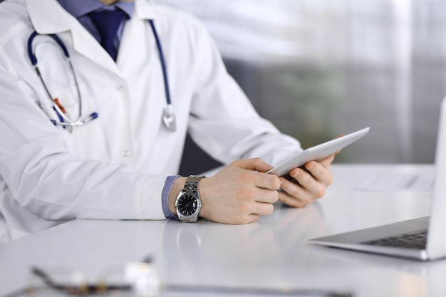 Unknown male doctor sitting and working with tablet computer in clinic at his working place, close-up. Young physician at work. Perfect medical service, medicine concept.