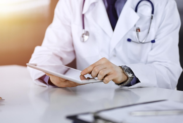 Unknown male doctor sitting and working with tablet computer iin a darkened clinic, glare of light on the background, close-up of hands