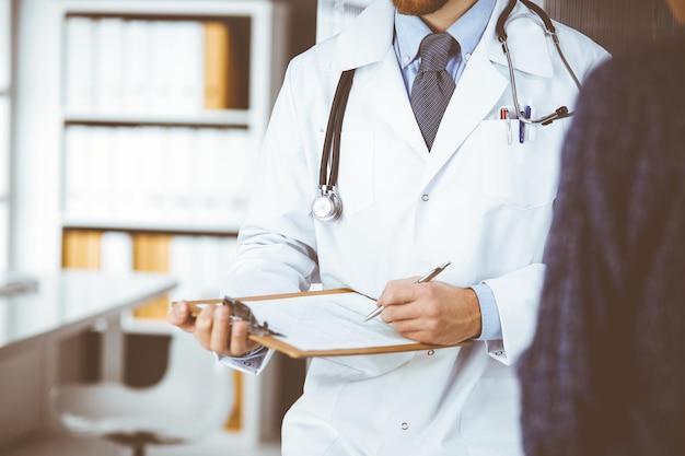 Unknown red-bearded doctor and patient woman discussing current health examination while sitting in sunny clinic, close-up. Medicine concept.