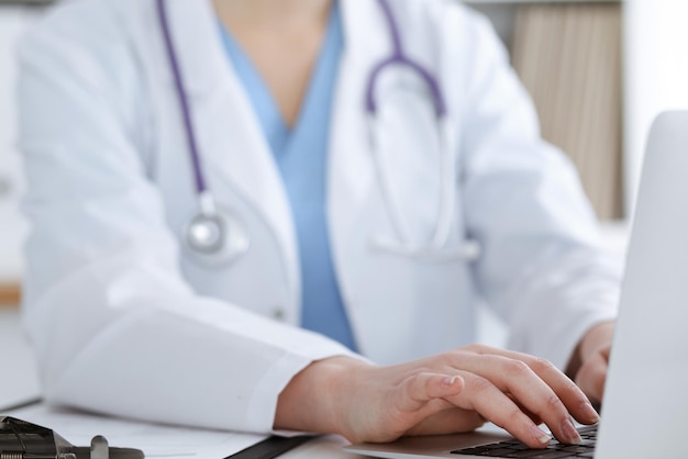 Unknown woman-doctor typing on laptop computer while sitting at the table. Medicine concept.