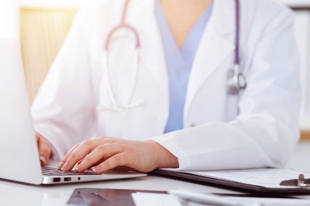 Unknown woman-doctor typing on laptop computer while sitting at the table in sunny clinic.