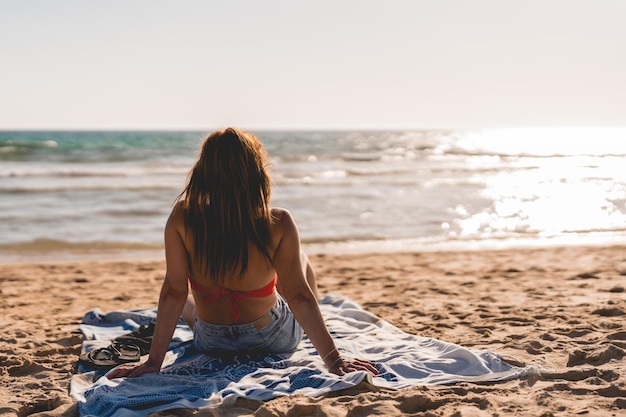 Unknown woman sitting on a towel on the beach watching the sea