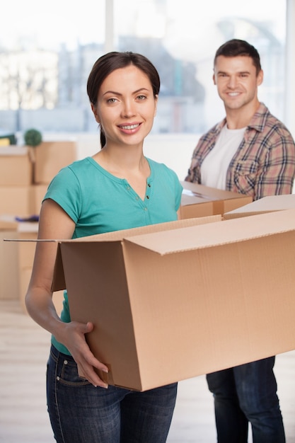 Unpacking boxes. Attractive young woman holding an opened cardboard box and smiling at camera while cheerful man standing on background