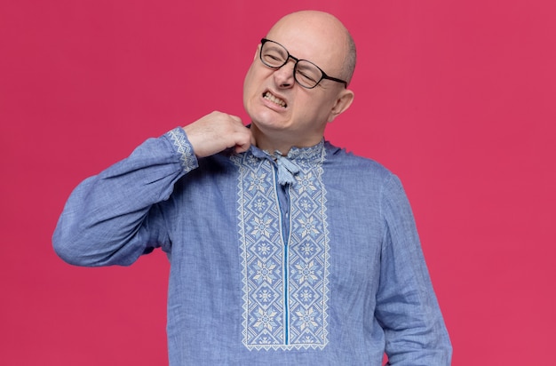 Unpleased adult slavic man wearing optical glasses pulling collar of his blue shirt