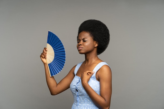 Unpleased afro woman suffer from heat inside overheated african female waving with paper fan