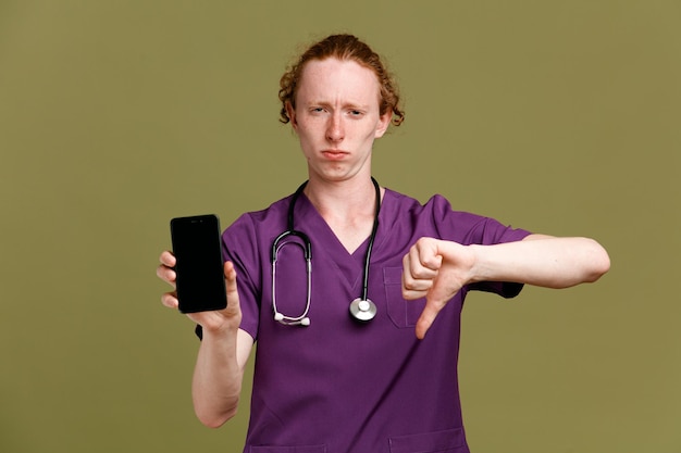 unpleased showing thumbs down holding phone young male doctor wearing uniform with stethoscope isolated on green background