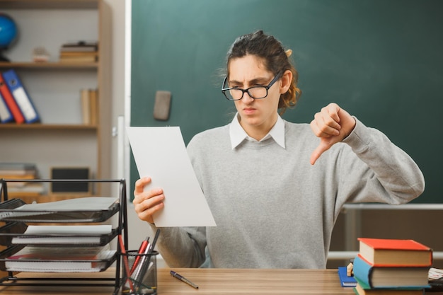 unpleased showing thumbs down young male teacher wearing glasses holding paper sitting at desk with school tools on in classroom