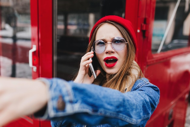 Unpleased stylish girl talking on phone near red bus Outdoor shot of emotional caucasian woman with bright makeup posing with smartphone in spring day