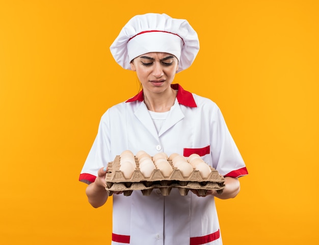 Unpleased young beautiful girl in chef uniform holding and looking at batch of eggs 