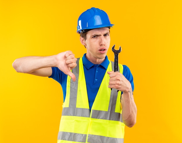 Unpleased young builder man in uniform holding open-end wrench showing thumb down isolated on yellow wall