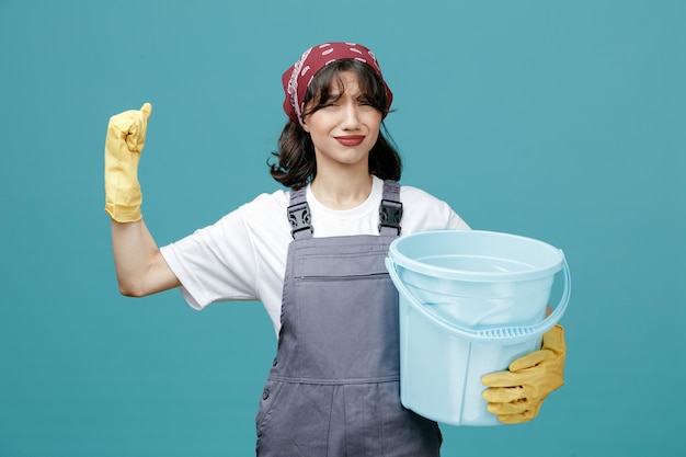 Unpleased young female cleaner wearing uniform bandana and rubber gloves holding bucket keeping fist in air looking at camera isolated on blue background