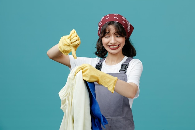 Unpleased young female cleaner wearing uniform bandana and rubber gloves holding dirty clothes looking at them showing thumb down isolated on blue background