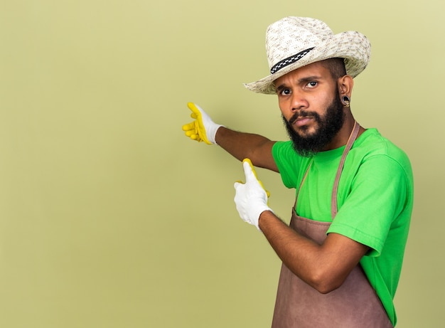 Unpleased young gardener afro-american guy wearing gardening hat with gloves points at behind 