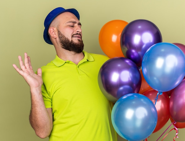 Unpleased young guy wearing party hat holding and looking at balloons spreading hand isolated on olive green wall