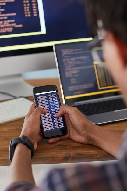 Photo unrecognizable african-american man holding smartphone with code on screen while working at desk in office, it developer concept, copy space