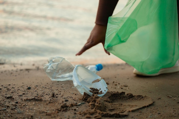 Unrecognizable African woman collecting spilled garbage from sand on beach in green plastic bag Ecology pollution