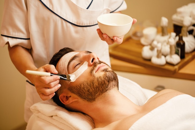 Unrecognizable beautician applying facial mask on man's face during spa treatment at wellness center