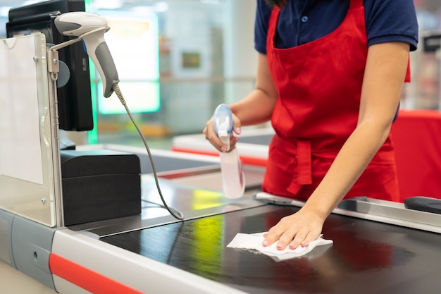 Unrecognizable cashier wearing red apron cleansing checkout surface with disinfectant in modern store