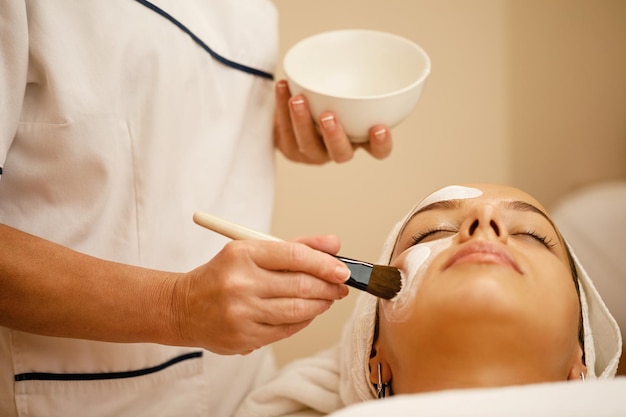 Unrecognizable cosmetician applying white detox mask on woman's face during facial treatment at beauty spa