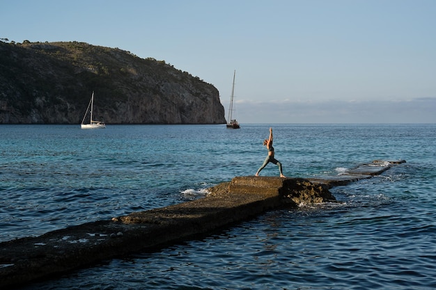 Unrecognizable female in sportswear practicing Crescent Lunge yoga on rocky cliff near rippling sea against sky in summer day