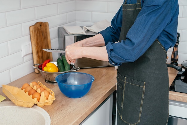 Unrecognizable man breaking egg with knife into blue plastic round bowl near steel colander with yellow bell pepper and wooden cutting board Cooking healthy food diet