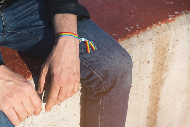 Unrecognizable man in jeans and black leather jacket sitting on a concrete bench with rainbow gay pride bracelet