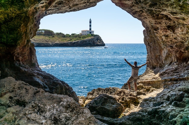 Unrecognizable man with raised hands inside a cave overlooking the sea and a red and white lighthouse with a ship sailing