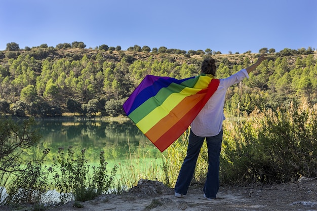 Unrecognizable middle-aged lesbian woman holding the Gay Rainbow Flag on at the lake outdoors. Freedom concept