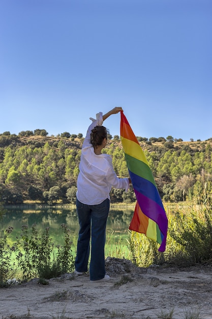 Unrecognizable middle-aged lesbian woman holding the Gay Rainbow Flag on at the lake outdoors. Freedom concept