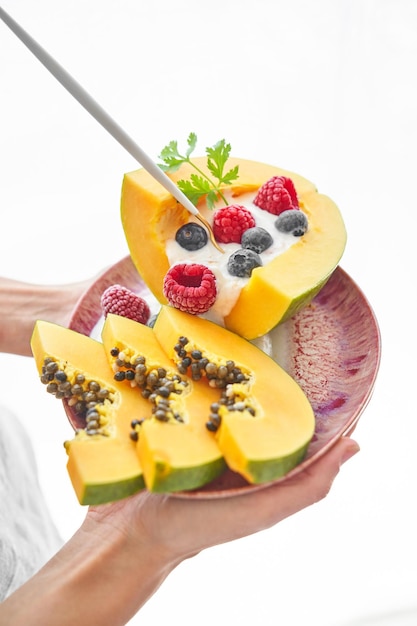 Unrecognizable person demonstrating portion of sweet dessert made of papaya raspberries blueberries and yogurt and served on pink ceramic plate with spoon