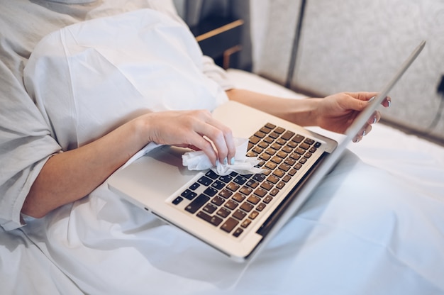 Unrecognizable woman cleaning laptop by hand sanitizer, using cotton wool with alcohol to wipe to avoid contaminating with COVID-19