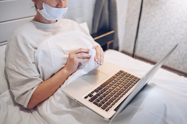 Unrecognizable woman in face mask in bedroom during coronavirus isolation home quarantine cleaning laptop by hand sanitizer, using cotton wool with alcohol to wipe to avoid contaminating with COVID-19