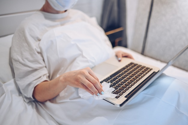 Unrecognizable woman in face mask in bedroom during coronavirus isolation home quarantine cleaning laptop by hand sanitizer, using cotton wool with alcohol to wipe to avoid contaminating with COVID-19