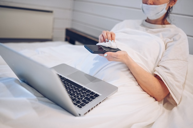 Unrecognizable woman in face mask in bedroom during coronavirus isolation home quarantine cleaning phone by hand sanitizer, using cotton wool with alcohol to wipe to avoid contaminating with COVID-19