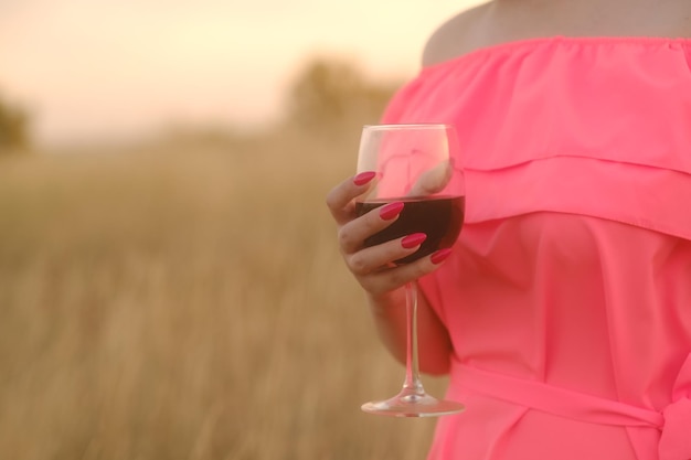 Unrecognizable woman holding glass of red wine standing in nature on sunset Body part of female in bright dress with alcohol in countryside